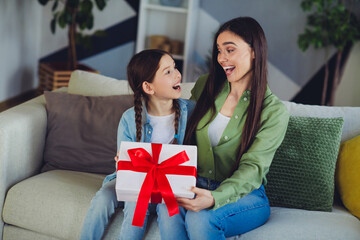 A joyful mother and daughter bonding moment while sharing a wrapped gift on a comfortable sofa in...