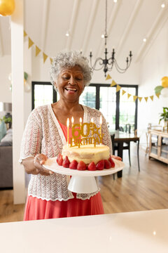 Senior African American woman holding 60th strawberry candlelit cake amid dining bunting balloons