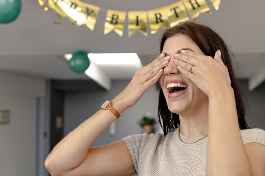 Woman covering eyes and laughing in party room with gold birthday banner green balloons, copy space