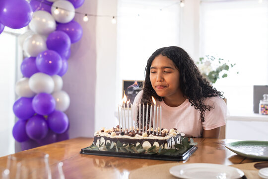 Child girl blowing out lit candles at wooden table with chocolate cake and balloon arch
