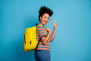 Young woman in colorful striped t-shirt with bright yellow backpack making a peace sign against a...