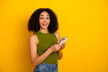 Young cheerful woman holding smartphone against yellow background, expressing joy and energy, ideal...