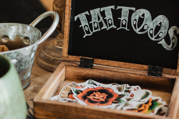 Rustic wooden box of vintage-style tattoo flash stickers beneath chalkboard “TATTOOS” sign on...