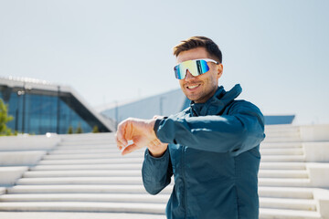 Smiling sports enthusiast checks smartwatch while standing on urban steps on a bright sunny day