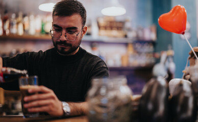 A bearded man wearing glasses is shown at a cozy lounge bar counter while pouring a drink, with warm lighting and a romantic ambience. A red heart-shaped balloon is in the foreground.