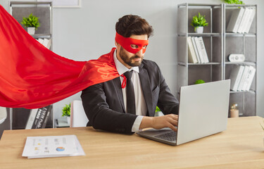 Serious man superhero wearing red mask and cape, working at laptop computer busy doing financial...
