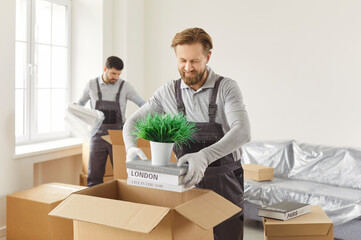 Bearded mover placing plant and travel guidebooks into open moving box while colleague unwrapping...