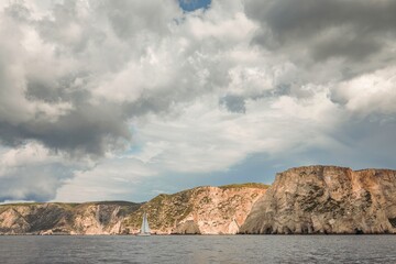 A lone sailboat glides gracefully across calm waters, framed by towering cliffs and a moody sky....