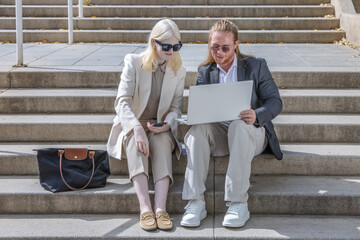 Business colleagues working on laptop outdoors on steps, albino woman and redhead man