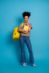 Young student in colorful attire holding book and wearing a yellow backpack posing joyfully against...