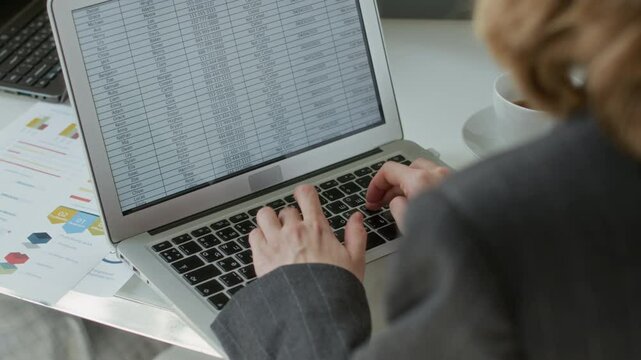 Over the shoulder view of businesswoman dressed in grey jacket working with spreadsheet displayed on screen of her laptop, documents and white coffee cup on white table