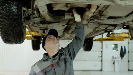 Professional mechanic wearing gloves carefully examining underside of vehicle lifted on hydraulic...