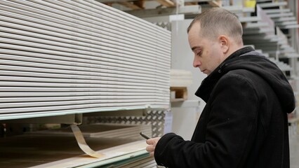 Obraz premium Construction professional reviewing building materials on smartphone screen, comparing drywall panels and plasterboard in warehouse storage area, selecting optimal supplies for project