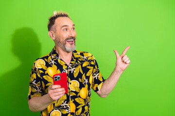 Happy mature bearded man in lemon print shirt holding red smartphone, gesturing excitement, on green background