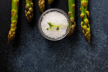 Fresh asparagus spears arranged alongside a bowl of coarse salt on a dark countertop surface