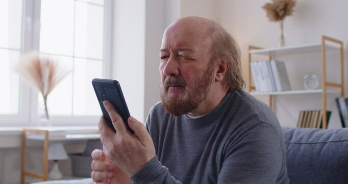 Worried elderly man holding smartphone and looking at screen with concern. Portrait of focused senior male sitting on sofa, having problems with modern mobile technology or receiving unexpected news.