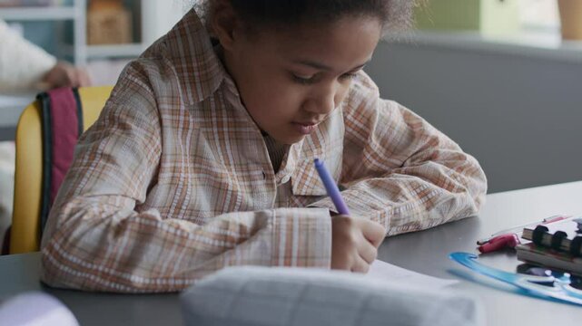 Zoom-in shot of young African American girl carefully reviewing her worksheet with pencil in hand and giggling, working quietly at her desk in structured classroom environment