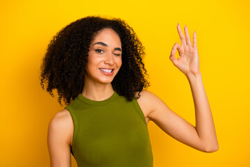 Smiling young woman winking and making an OK hand gesture against a vibrant yellow background in...