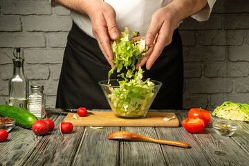 A chef mixes fresh greens and vegetables in a glass bowl on a wooden surface. Bright tomatoes and...