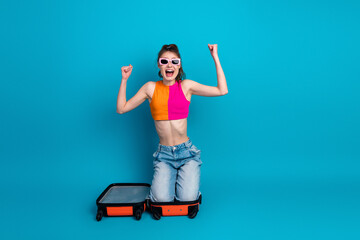 Excited young woman sitting inside a suitcase with cheerful expression and fashionable casual...