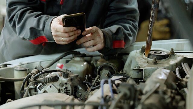 Car mechanic checking vehicle engine using smartphone, displaying digital diagnostic technology in automotive maintenance
