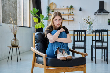 Portrait of young beautiful serious woman looking at camera sitting at home in chair