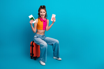 Young lady seated on a suitcase while holding a phone and passport against a vibrant cyan background