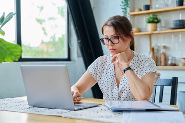 Middle aged serious woman working with laptop sitting at table at home