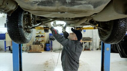 Experienced mechanic checking vehicle undercarriage with flashlight, working carefully in professional automotive repair workshop, wearing protective gloves