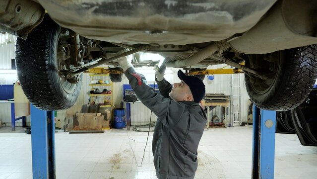 Experienced mechanic checking vehicle undercarriage with flashlight, working carefully in professional automotive repair workshop, wearing protective gloves