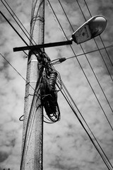 A weathered utility pole stands against a bright blue sky, entangled with a chaotic mess of black...