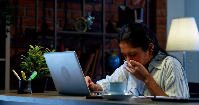 Businesswoman having cough and cold while working on laptop at desk in modern cozy office, Indian Asian young female employee using tissue to sneeze while showing symptoms of flu, physical discomfort