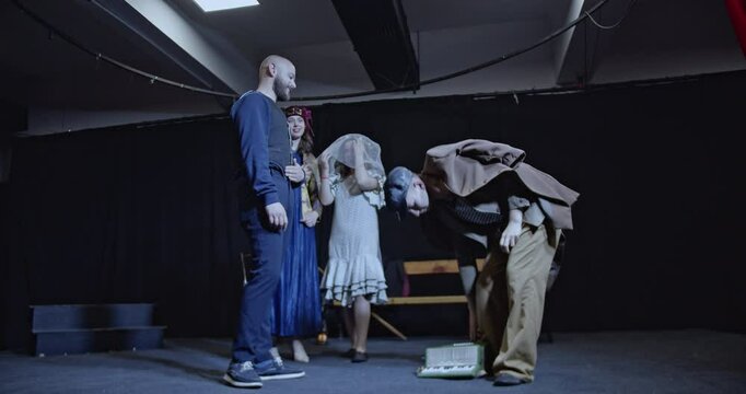 Young actors rehearse a play on a theater stage. A man plays the accordion while others act. The actors prepare for their roles.