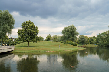 Green hill by the calm lake with cloudy sky and trees in a public park