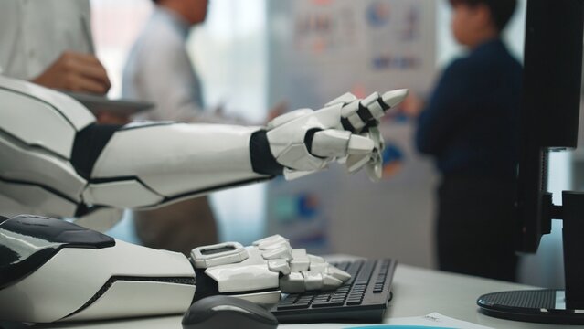 A robotic hand extends towards a keyboard in a modern office. Professionals engage in discussions, highlighting innovation and technology in a collaborative setting.Noogenesis