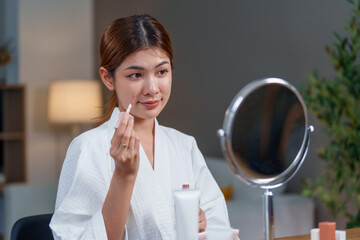 Woman applies makeup in front of mirror after her shower, focusing on skin care to enhance her...