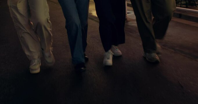 A group of cheerful young people are walking around the city at night with their arms around each other. They're talking. The video moves from close-up of their legs to their faces