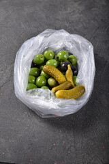 Green olives, black olives and pickled gherkins in a cellophane bag on a gray background