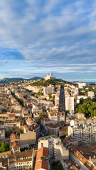 Drone aerial view of the city of Marseille, France, with the iconic Notre-Dame de la Garde basilica on a hilltop surrounded by urban buildings under a partly cloudy sky.