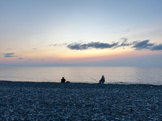 Fototapeta premium Two people fishing on pebble beach at sunset, peaceful sea view