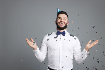 Happy Birthday. Handsome man with party hat under confetti shower on grey background, space for text