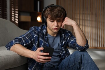 Obraz premium Teen problems. Upset teenage boy in headphones with smartphone near sofa at home