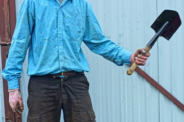 one small short compact wooden handle industrial sharp iron black dangerous military army sapper shovel in hand in glove of man in blue shirt and pants day outside