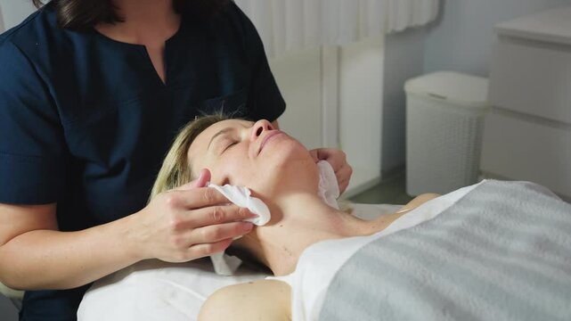 Under bright lamp light massage therapist gently cleanses face of female client with damp cotton pad before skin care treatment in modern white clinic room with calm relaxing atmosphere