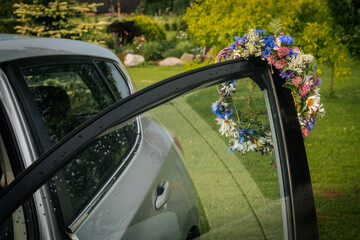A wreath of wildflowers hangs on the edge of an open car door, raindrops fall on the glass, and in the background - a lush green garden.