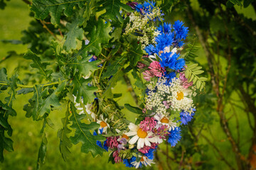 A close-up of a handmade wreath made of wildflowers and oak leaves hanging outdoors in a green...