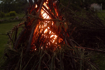 A dark, moody image of a bonfire made of thick branches and adorned with a wreath of braided grain,...