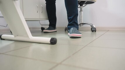 Leg view of treatment provider wearing crocs and navy blue uniform standing beside clinical bed under bright lamp with mobile stool, product table and cabinet visible on clean tiled floor