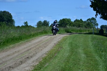 Fototapeta premium Motorcyclist rides an adventure touring motorcycle on a gravel countryside road. Rural landscape with green grass and blue sky. Concept of freedom, summer trip, and rural travel lifestyle.