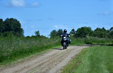 Motorcyclist rides an adventure touring motorcycle on a gravel countryside road. Rural landscape with green grass and blue sky. Concept of freedom, summer trip, and rural travel lifestyle.